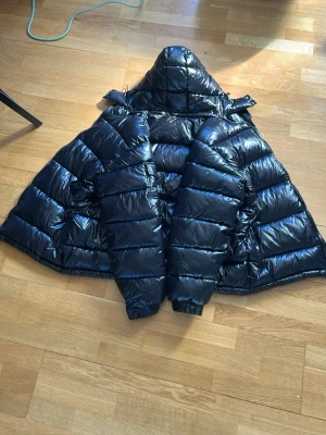 Svart dunjacka från Moncler - Säljer en svart, glansig dunjacka från Moncler med huva och breda, quiltade kanaler. Jackan har en klassisk puffer-look och är perfekt för kalla vinterdagar. Stängs med dragkedja och har detaljer i metall. Riktigt snygg och trendig vinterjacka.