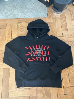 Maison Margiela hoodie - Svart hoodie från Maison Margiela med röd och vit grafisk print framtill. Klassisk huva och känguruficka. Snygg och modern design med märkets ikoniska siffror och logga. De vita prickarna man ser i bilden är bara dam som tas bort när vi skickar.