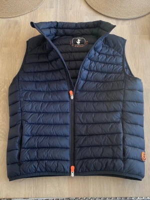 Mörkblå dunväst från Anima - Snygg mörkblå dunväst från Anima med hög krage och orange detaljer på dragkedjan. Västen har quiltad design, två sidofickor och ett runt brunt logomärke på nederkanten. Perfekt för lager-på-lager och riktigt skön att bära.