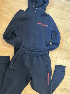 Svart set från Prada med huva - Snyggt svart set från Prada med matchande hoodie och byxor. Hoodien har dragkedja och röd logodetalj på bröstet, byxorna har resår i midjan, dragkedjefickor och röd logodetalj på benet. Perfekt för en avslappnad och trendig look. ‼️en dragskedja till fickan har gått av på byxorna och som jag visar finns en liten fläck inuti tröjan😌 noppror finns även på vissa ställen‼️