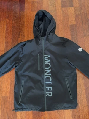 Svart vindjacka från Moncler - Lätt svart Moncler Zind-jacka med huva och en stor vertikal 'MONCLER'-logotyp framtill. Denna modell har en hel dragkedja och en Moncler-logotyplapp på vänster ärm. Jackan är i utmärkt skick, utan synliga tecken på slitage, och den inre etiketten indikerar att den är fylld med vitt gåsdun.  ⭐️ Skick: 9/10 🎨 Färg: Svart 📦 Snabb och noggrann leverans