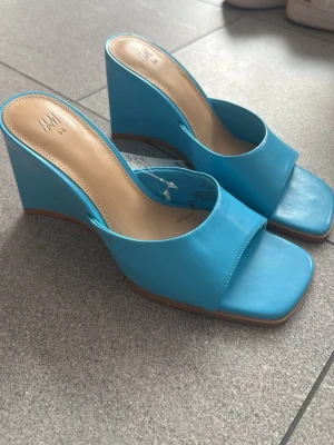 Blåa wedge-sandaler från H&M - Snygga blåa sandaler med kilklack från H&M. De har öppen tå, bred rem över foten och fyrkantig tåform. Tillverkade i konstläder med beige innersula och bruna kanter. Perfekta för att lyfta din outfit med en pop av färg.