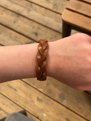 Flätat brunt läderarmband - Säljer ett snyggt flätat armband i brunt läder med justerbar knäppning med tre metalltryckknappar. Perfekt för dig som gillar en enkel och stilren look. Armbandet har en klassisk design och passar till många olika outfits.