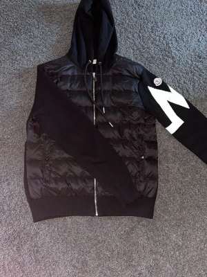 Svart pufferjacka med huva från Moncler - Cool svart pufferjacka från Moncler med stickade ärmar och stor huva. Jackan har dragkedja framtill, två fickor och ett vitt zigzag-mönster på ena ärmen samt Moncler-logga. Perfekt för kalla dagar och snygg streetstyle. Storleken är M men passar en S