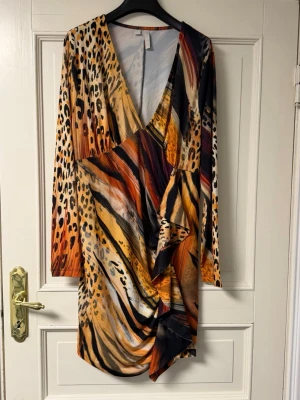 Zebramönstrad omlottklänning - Cool kort omlottklänning med v-ringning och lång ärm. Klänningen har ett djuriskt zebramönster i svart, orange, beige och brunt. Tillverkad i återvunnen polyester, perfekt för dig som gillar att sticka ut.