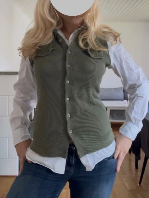 Olivgrön ribbad väst med knappar - Snygg olivgrön väst med ribbad struktur och knäppning framtill. Västen har två bröstfickor med lock och en klassisk krage. Perfekt att styla över en skjorta för en trendig lager-på-lager-look.