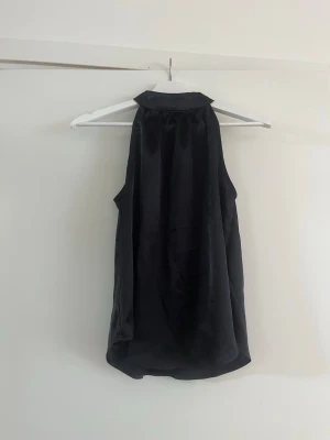 Svart ärmlös satinlinnetopp - Stilren svart linnetopp i glansigt satinmaterial med hög krage och ärmlös design. Perfekt att matcha med jeans eller kjol för en trendig look. Toppen har en lös passform och faller snyggt över kroppen. Vet ej märket då lapparna är bortklippta. Aldrig använd. 
