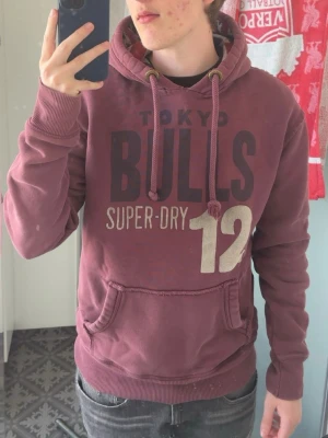 Mörkröd hoodie från Superdry med tryck - Säljer en mörkröd hoodie från Superdry med stort tryck på bröstet och känguruficka. Och även snygg fodring vid luvan