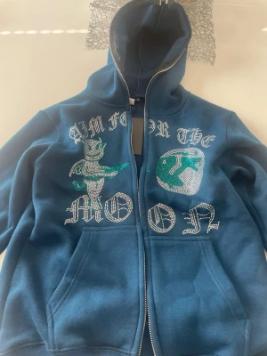 Blå hoodie med tryck och dragkedja - Snygg blå hoodie med dragkedja och huva. Framsidan har ett stort tryck med texten 'I'M FOR THE MOON', en tecknad figur och en jordglob i grönt och vitt. Hoodien har kängurufickor och är i mjukt material, perfekt för en chill stil.