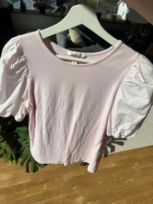 Ljusrosa topp med puffärmar - Supersöt ljusrosa topp med korta puffärmar i vitt. Toppen har rund halsringning och är gjord i mjukt bomullstyg. Perfekt för dig som vill ha en feminin och trendig look med extra volym på ärmarna.