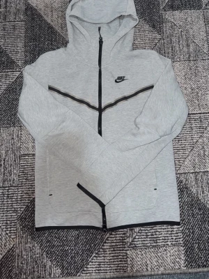 Ljusgrå Nike hoodie med dragkedja - Snygg ljusgrå hoodie från Nike med hel dragkedja och svart Nike-logga på bröstet. Hoodien har svarta detaljer längs bröstet och två fickor framtill. Perfekt för en sportig och avslappnad stil. Tillverkad i mjukt och bekvämt material.