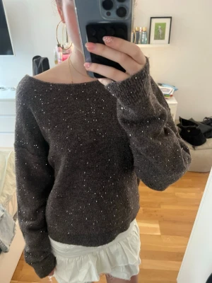 Glittrig brun stickad tröja med öppen rygg - Mysig stickad tröja i brun färg med glittriga paljetter. Tröjan har en bred, off-shoulder halsringning och en cool öppen rygg med en dekorativ rem. Perfekt för att sticka ut och ge en extra touch till din outfit.