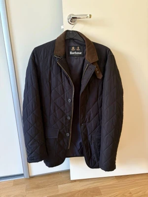 Svart quiltad jacka från Barbour - Snygg svart quiltad jacka från Barbour med brun krage i manchester. Jackan har dragkedja och knappar framtill, två fickor och klassiskt rutmönster. Perfekt för dig som vill ha en stilren och tidlös look.