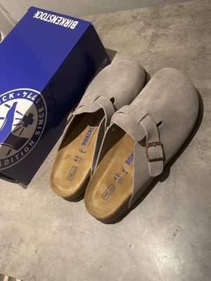 Birkenstock Boston tofflor mocka grå - Säljer ett par Birkenstock Boston tofflor i grå mocka med justerbart spänne i metall. Klassisk slip-in modell med korksula och mjuk fotbädd för extra komfort. Perfekta för dig som vill ha både stil och bekvämlighet. Passar även 44