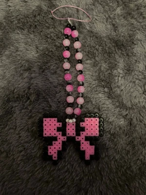 Rosa pärlaccessoar med pixelrosett - Unik accessoar med pärlor i rosa och svart, avslutas med en pixelinspirerad rosett i rosa och svart plast. Perfekt att hänga på väskan eller använda som nyckelring. Handgjord och färgglad med retrovibe.