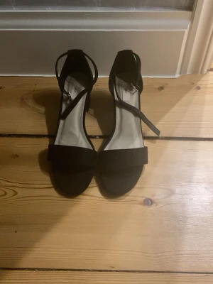 Svarta klackskor med rem.  - Säljer ett par svarta låga klackskor och justerbar ankelrem med silverfärgat spänne. Skorna har öppen tå och bred rem över foten. Klassisk och stilren design som passar till många olika outfits.