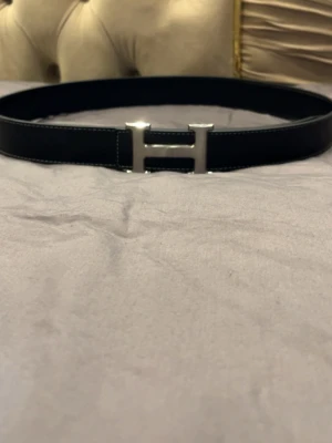 Svart skärp med H spänne - Snyggt svart skärp med H spänne. Skärpet har stilrena vita sömmar längs kanterna. Perfekt accessoar för att lyfta din outfit.