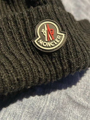 Svart ribbstickad mössa från Moncler - Snygg svart ribbstickad mössa från Moncler med klassisk logotyp framtill. Mössan har uppvikt kant och är tillverkad i ett mjukt och värmande material som passar perfekt för kyliga dagar.