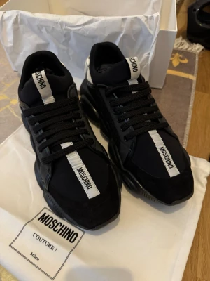 Äkta Moschino sneakers med logga - Snygga svarta sneakers från Moschino med tydlig vit logga på både plös och häl. Skorna har en chunky sula, svarta skosnören och detaljer i mocka och textil. Den vita remmen med Moschino-tryck ger en extra cool touch. Perfekta för dig som gillar statement-skor.