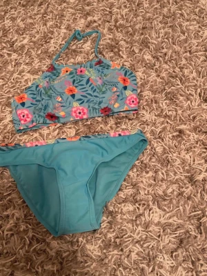 Blommig blå bikini med hög hals - Snygg bikini med blommigt mönster i rosa, orange och rött på blå botten. Överdelen har hög hals och smala axelband som knyts i nacken. Underdelen är enkel i blått med blommig kant upptill. Perfekt för strandhäng och poolparty!