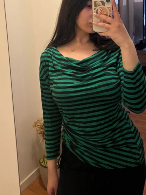 Randig topp från Michael Kors - Snygg långärmad topp från Michael Kors i grönt och svart med randigt mönster. Toppen har en lösare passform och en draperad, vid ringning som ger en cool och avslappnad look. Perfekt att styla med jeans eller kjol.