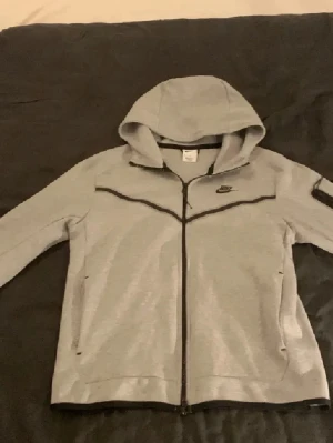 Grå Nike hoodie med dragkedja -  grå hoodie från Nike med hel dragkedja och svart Nike-logga på bröstet. Hoodien har svarta detaljer längs dragkedjan och bröstet samt praktisk huva. Aldrig använts så inga defekter