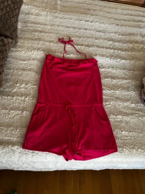Rosa kort jumpsuit med knytband - Snygg rosa jumpsuit med halterneck och knytband i nacken. Modellen är kort med shorts och har resår i midjan samt fickor på sidorna. Perfekt för varma dagar och enkel att styla med sneakers eller sandaler.