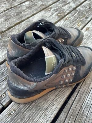 Valentino Garavani sneakers med nitar - Snygga sneakers från Valentino Garavani i svart och brunt läder med coola nitar på sidorna. Skorna har svarta snören, svart meshfoder och beige sula med nitar på hälen. Perfekta för dig som gillar en edgy och unik stil.