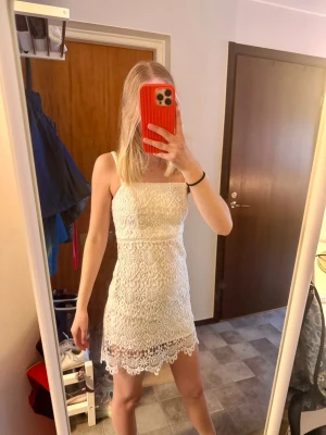 Vit spetsklänning med smala axelband, studentklänning - Säljer en vit, kort klänning i spets med smala axelband och rak urringning. Klänningen har ett figurnära snitt och fina virkade spetsdetaljer längs fållen. Perfekt för sommarens alla tillfällen och ger en romantisk vibe.