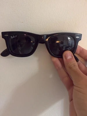 Ray-Ban glasögon - Säljer ett par klassiska svarta Ray-Ban Wayfarer solglasögon med mörka linser och ikonisk logga på linsen. Snygg och tidlös design som passar till många olika stilar. Bågarna är gjorda i syntetmaterial.