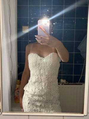 Kort Vit spetsklänning axelbandslös - Säljer en vit, axelbandslös klänning med vacker spets och blommigt mönster. Klänningen har en figurnära passform och är kort i modellen, perfekt för sommarens alla tillfällen. Supersnygg och trendig design!