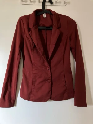 Mörkröd figursydd kavaj röd brun - Snygg mörkröd kavaj med figurnära passform och klassisk krage. Kavajen har två knappar framtill och långa ärmar. Perfekt för en stilren look. Materialet känns mjukt och följsamt.