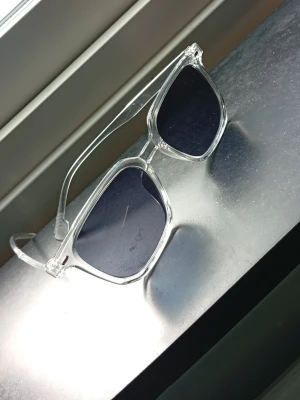 Solglasögon  - Snygga solglasögon med transparent plastbåge och mörka, fyrkantiga glas. Ramen är helt genomskinlig vilket ger en modern och clean look. Perfekta för dig som vill ha en stilren accessoar till sommaren.