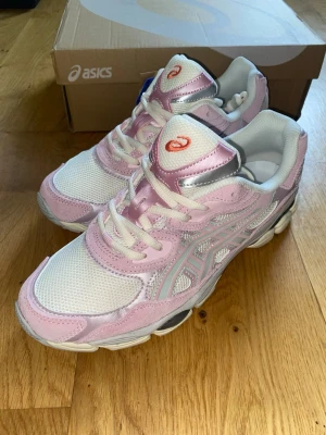Asics GEL-NYC - Snygga ASICS sneakers i vitt och ljusrosa med silvriga detaljer. Ovandelen är i mesh och syntetmaterial för en sportig look. Klassisk snörning och låg profil gör dem perfekta till streetwear. ASICS-logga på plösen och sidan.