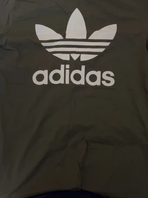Svart Adidas t-shirt med logga - Cool svart t-shirt från Adidas med stor vit Trefoil-logga och text på bröstet. Klassisk passform och rund halsringning, perfekt för en sportig och avslappnad stil.