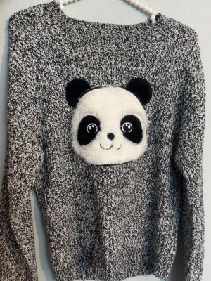 Gråmelerad stickad tröja med panda - Mysig gråmelerad stickad tröja med lång ärm och en stor, fluffig pandadekor framtill. Tröjan har rund halsringning och ribbstickade muddar. Perfekt för dig som gillar gulliga detaljer och vill sticka ut lite extra.