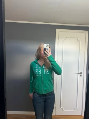Grön hoodie från Hollister - Snygg grön zip up hoodie från Hollister med vit text på bröstet. Tröjan har dragkedja, två fickor fram och en huva med snören. Materialet är mjukt och bekvämt.