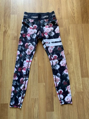 Blommiga leggings från Stronger - Supersnygga leggings från Stronger med svart bas och blommigt mönster i rosa och vitt. Hög midja med logotyp runt om och vita ränder på ena benet. Perfekta för träning eller chill. Stretchigt material som sitter skönt.