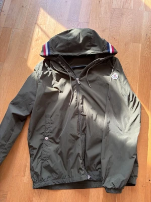 Monclers Olivgröna Windbreaker - Snygg och skön olivgrön windbreaker från Moncler med huva och dragkedja framtill. Jackan har monclers logga på ena armen och en smidig och välfungerande dragkedja. Luvan fungerar toppen då man kan även ändra passform med snörena! Jackan är som perfekt nu till våren, med lite blåst även småkyliga dagar. Några övriga frågor, kontakta mig! Storlek 3 passar S och M skriv vid minsta fundering🙌🏻🙌🏻