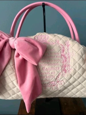 Juicy Couture quiltad handväska rosa rosett - Supersöt quiltad handväska från Juicy Couture i ljusbeige sammet med rosa detaljer, broderad logga och stor rosa rosett. Väskan har två rosa handtag och romantisk vibe, perfekt för dig som gillar gulliga accessoarer.