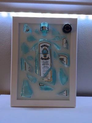 Tavla med krossad Bombay Sapphire-flaska - Unik tavla med en krossad ljusblå Bombay Sapphire gin-flaska arrangerad i en vit ram. Flaskbitarna och etiketten är konstnärligt placerade för en cool och modern look. Perfekt som dekoration för hemmabaren eller köket. Tavlan är egengjord av Epoxycreations och är gjord av återbruk!🤩
