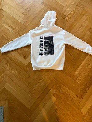 Vit hoodie med tryck 'Believe' - Snygg vit hoodie med huva och känguruficka. På bröstet står det 'Believe' i svart text och på ryggen finns ett stort tryck med texten 'Believe' och en svartvit bild. Perfekt för en cool och avslappnad stil.
