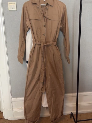 Beige utility jumpsuit med bälte - Stilren beige utility jumpsuit med långa ärmar, knappar framtill och två bröstfickor. Tillhörande midjebälte för snygg passform och klassisk krage. Perfekt för en trendig och avslappnad look. Materialet känns lätt och bekvämt.