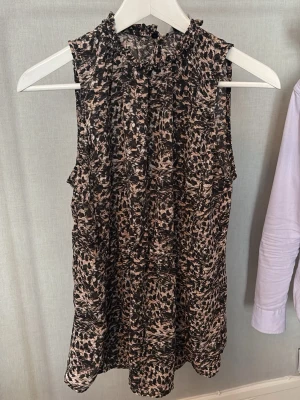 Leopardmönstrad ärmlös blus - Säljer en snygg ärmlös blus med leopardmönster i svart och beige. Blusen har hög krage med små volangdetaljer och ett luftigt, lätt tyg som ger en skön känsla. Perfekt för dig som vill sticka ut lite extra.