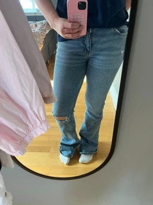 Blå bootcut jeans - Säljer ett par ljusblå bootcut jeans med slitning och hål på ena knät. Jeansen har klassisk femficksdesign och hög midja. Perfekta för en avslappnad och trendig look. Materialet är denim i bomull.