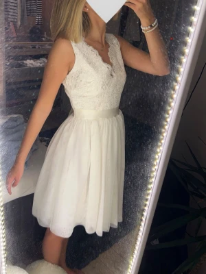 Vit spetsklänning med v-ringning, Studentklänning  - Söt vit klänning med v-ringad spets upptill och markerad midja med satinband. Kjolen är lätt utsvängd och slutar ovanför knät. Ärmlös modell som ger en elegant och feminin look. Perfekt för student eller sommarens speciella tillfällen 🎀