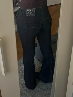Svarta bootcut jeans True Religion - Säljer ett par svarta bootcut jeans från True Religion med låg midja och snygga fickdetaljer med knappar. Jeansen är snyggt uppsydda och passar mig som är 164 cm lång bra. Storlek 24. Tveka inte att skriva vid frågor💕
