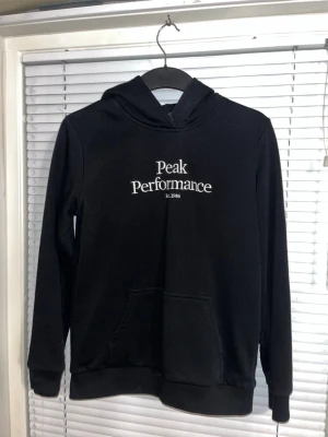 Svart Peak Performance hoodie - Snygg svart hoodie från Peak Performance med vit broderad logga på bröstet. Den är i jättebra skick. Jag fick den men har aldrig använt den. Basic hoodie som man kan ha till alla outfits. 