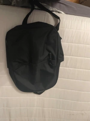 Svart enkel ryggsäck - Säljer en stilren svart ryggsäck med minimalistisk design. Väskan har ett stort huvudfack och en ytterficka framtill. Tillverkad i slitstarkt tygmaterial och har justerbara axelremmar. Perfekt för skola eller cityhäng. Den är 30x40