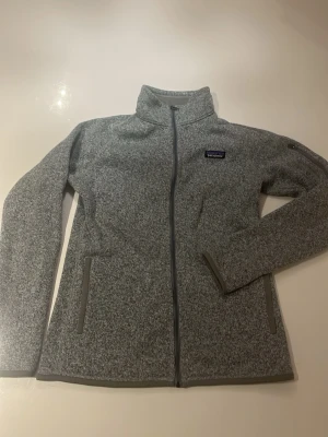 Grå fleecejacka från Patagonia - Snygg gråmelerad fleecejacka från Patagonia med hel dragkedja framtill, två sidofickor med dragkedja och hög krage. Jackan har en liten logotyp på bröstet och är perfekt för lager-på-lager-stil under kyliga dagar.